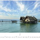 Unterer Text: Überlingen Pfahlbauten. Oben Pfahlhäuser über Wasser, rechts Bäume, blauer Himmel mit Wolken.