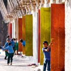Keine Texte. Kinder spielen zwischen bunten Fensterläden unter einem Dach aus Bambus. Sandalen liegen am Boden.