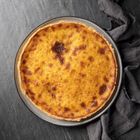 Ein runder, goldbrauner Kuchen auf einem grauen Teller liegt auf dunklem Stoff und einer steinartigen Oberfläche.