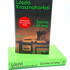 Vorderseite eines Buches: "László Krasznahorkai, Zsömle ist weg, Nobelpreis für Literatur 2025". Oranges Cover mit Kaffeetisch-Illustration.