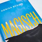 Der Text enthält "Annika Büsing" und "MAGISCH" in großen gelben Buchstaben auf blauem Hintergrund mit schwarzem Hügelmotiv.
