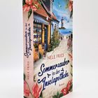 „Sommerzauber in der Inselapotheke“ von Nele Fries. Eine idyllische Küstendorf-Szene mit Haus, Blumen und einem Leuchtturm.