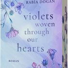 Titel: „violets woven through our hearts“. Autor: RABİA DOĞAN. Mit violetten Blumen auf pastellfarbenem Hintergrund.
