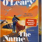 „Wer ist hier eigentlich wer?“ Oben der Name Beth O'Leary. Illustration: Paar am Rand einer Klippe, Leuchtturm im Hintergrund.