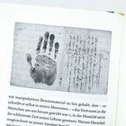 Links ein großer schwarzer Handabdruck. Rechts handschriftliche Notizen; unleserlich und schräg verlaufend. Darunter gedruckter Text.