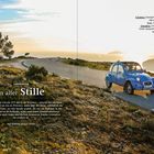 FRANKREICH; In aller Stille. Ein blauer Citroën 2CV fährt auf einer kurvigen Straße bei Sonnenuntergang durch eine hügelige Landschaft.