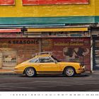 "4 SEASONS" steht groß auf einem Rolltor. Darunter ein gelbes, klassisches Auto. Gebäudefassade in Gelb und Grün.