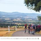 Ein Kalender zeigt Februar unten. Radfahrer fahren auf einer Landstraße. Die Landschaft ist hügelig mit Feldern und Bergen.