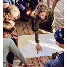 Unten steht "02" und die Tage des Monats. Fünf Personen betrachten interessiert eine ausgebreitete Seekarte auf einem Holzdeck.