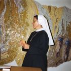 Eine Nonne steht vor einem Mosaik. Daneben sitzt eine Frau in einem geblümten Oberteil.