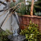 Ein runder Metallbehälter mit rauchendem Inhalt, umgeben von Tannenzweigen und Mistelzweigen, vor einem Holzrad.