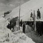 Mehrere Menschen räumen Schnee vor einem Gebäude auf einem verschneiten Hang. Sie tragen Winterkleidung.
