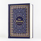 "Der Koran" auf einem kunstvoll verzierten, dunkelblauen Buch mit goldenen Ornamenten und detailreichem Muster.