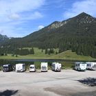 Kein sichtbarer Text. Wohnmobile stehen am Ufer eines Sees mit bewaldeten Bergen im Hintergrund.