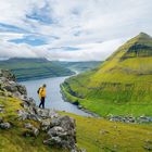 Keine Texte vorhanden. Eine Person in gelber Jacke steht auf einem grasbewachsenen Hügel, mit Blick auf ein Tal und eine Bucht.
