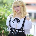 Junge Person mit blonden Haaren, trägt ein weißes Oberteil und schwarzen Rock. Goldene Schlüssel-Halskette. Grüne Kulisse.