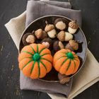 Zwei Filzkürbisse und Eicheln auf einem Teller, platziert auf gefalteten Stoffen in Erdtönen auf dunklem Untergrund.