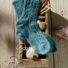 Zwei gestrickte, grüne Socken, dekorativ auf Holzkiste mit Garnknäueln und Stricknadeln arrangiert. Holzoptik im Hintergrund.