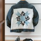 Jacke aus Jeansstoff mit einem großen, blumenbedruckten Patchwork-Muster auf dem Rücken. Darunter glänzende Schuhe.