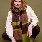 Ein lächelnde Person in einer weißen Jacke, fliederfarbenen Oberteil, braun-grün gestreiftem Schal, kniet auf olivgrünem Hintergrund.