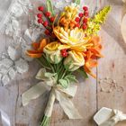 Häkelblumenstrauß in Orange und Gelb auf Holz, mit weißem Schleifenband, Spitze, Sandalen und einem Perlenarmband.