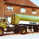Auf einem Tankwagen steht "HANIEL HEIZÖL". Der LKW ist grün und steht vor einem Backsteingebäude.