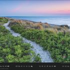 „02“, darunter Zahlen vom 01 bis 28. Ein sandiger Pfad führt durch grüne Dünen zum Meer unter einem rosa-blauen Himmel.