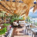 Terrasse eines Restaurants mit Holzboden, Pflanzen, Korbmöbeln und dekorativen Lichtern. Blick auf eine Bucht im Hintergrund.