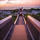 Februar 2027 in Lila. Ein hölzerner Weg führt über Dünen zum Meer bei Sonnenuntergang, mit teils bewölktem Himmel.