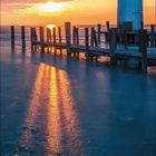 Kalenderseite für Februar 2027. Ein Leuchtturm bei Sonnenuntergang, reflektiert im Wasser, Holzpfähle führen zum Horizont.