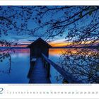 Kalender: Monat 2, beginnt mit Montag. Holzhütte auf Steg, umgeben von Wasser und Ästen bei Sonnenuntergang.