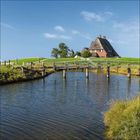 Ein Reetdachhaus liegt auf einem Hügel, umgeben von grünen Wiesen. Eine Holzbrücke führt über einen Wasserlauf.