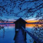 Keine erkennbaren Texte. Ein Steg führt zu einer Hütte über einem ruhig spiegelnden See bei Sonnenuntergang. Zweige im Vordergrund.