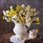 Gelbe und weiße Blumen in einer weißen Vase auf einem Holztisch mit Spitze. Daneben eine kleine Tasse und Blumen.