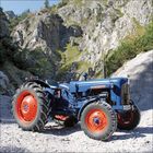 Ein blauer Traktor mit orangen Rädern steht vor einer felsigen Landschaft mit grüner Vegetation.