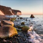Kein Text. Küstenlandschaft mit großen Felsen am Ufer, ruhiges Wasser und Klippen im Hintergrund bei Sonnenuntergang.