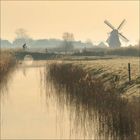 Ein Radfahrer auf einer Brücke über einem Kanal, im Hintergrund eine Windmühle und neblige Landschaft.
