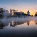 Eine Burg und ein Kirchturm spiegeln sich im nebligen Wasser bei Sonnenuntergang.