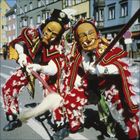 Karnevalsteilnehmer in roten, mit Federn dekorierten Kostümen und Holzmasken, halten Besenstiele; Hintergrund bunte Häuser.