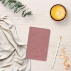 2027 auf einem rosa Notizbuch mit Blumenmuster, daneben eine brennende Kerze, Blattpflanze, Bleistift und Stoff.