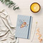 Ein blau-weiß gestreiftes Notizbuch mit einem Boot-Logo, umgeben von einem Creme-Stoff, Pflanze, Kerze und gestreiften Stift.