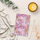 Ein Notizbuch mit floralem Muster, Stift, Kerze, Eukalyptusblätter und Stoff auf hellem Holztisch arrangiert.