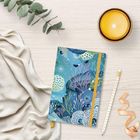 Ein Notizbuch mit Meeresmotiv, eine brennende Kerze, ein Bleistift, ein Band und Eukalyptus auf hellem Holz.