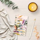 Notizbuch mit der Zahl "2027" und farbigen Blättern. Daneben ein Bleistift, Band, gelbe Kerze, Stoff und Eukalyptuszweig.