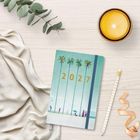 "2027" steht auf einem Tagebuch mit Palmenmotiv. Daneben liegen eine Kerze, ein Stift, Stoff, ein Band, Zweige.