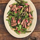 Ein ovaler Teller mit gebratenen Fleischscheiben, grünem Gemüse und Radieschen, auf einem Holztisch serviert.