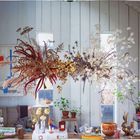 Text: "AUSSTELLUNG" und andere unlesbare Wörter. Trockenblumenarrangement schwebt in einem hellen, gemütlichen Raum.