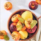 Februar. Holzteller mit Kakis, Birnen und roten Früchten auf hellem Hintergrund. Einige Früchte liegen daneben.