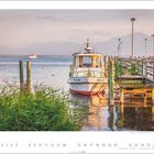 „Ingrid“ steht auf einem Boot, das an einem Steg vertäut ist. Dahinter ein weiter See mit Bergen im Hintergrund.