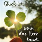 "Glück ist, wenn das Herz tanzt." Ein Sonnenstrahl hinter einem vierblättrigen Kleeblatt, gehalten von Hand.